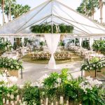Clear top tent reception setup with draped fabric, cross-back chairs, lush greenery, and white floral arrangements