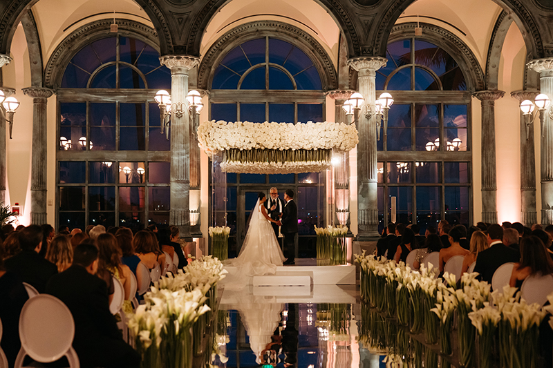 Grand ballroom ceremony with white floral chuppah and arched windows at twilight