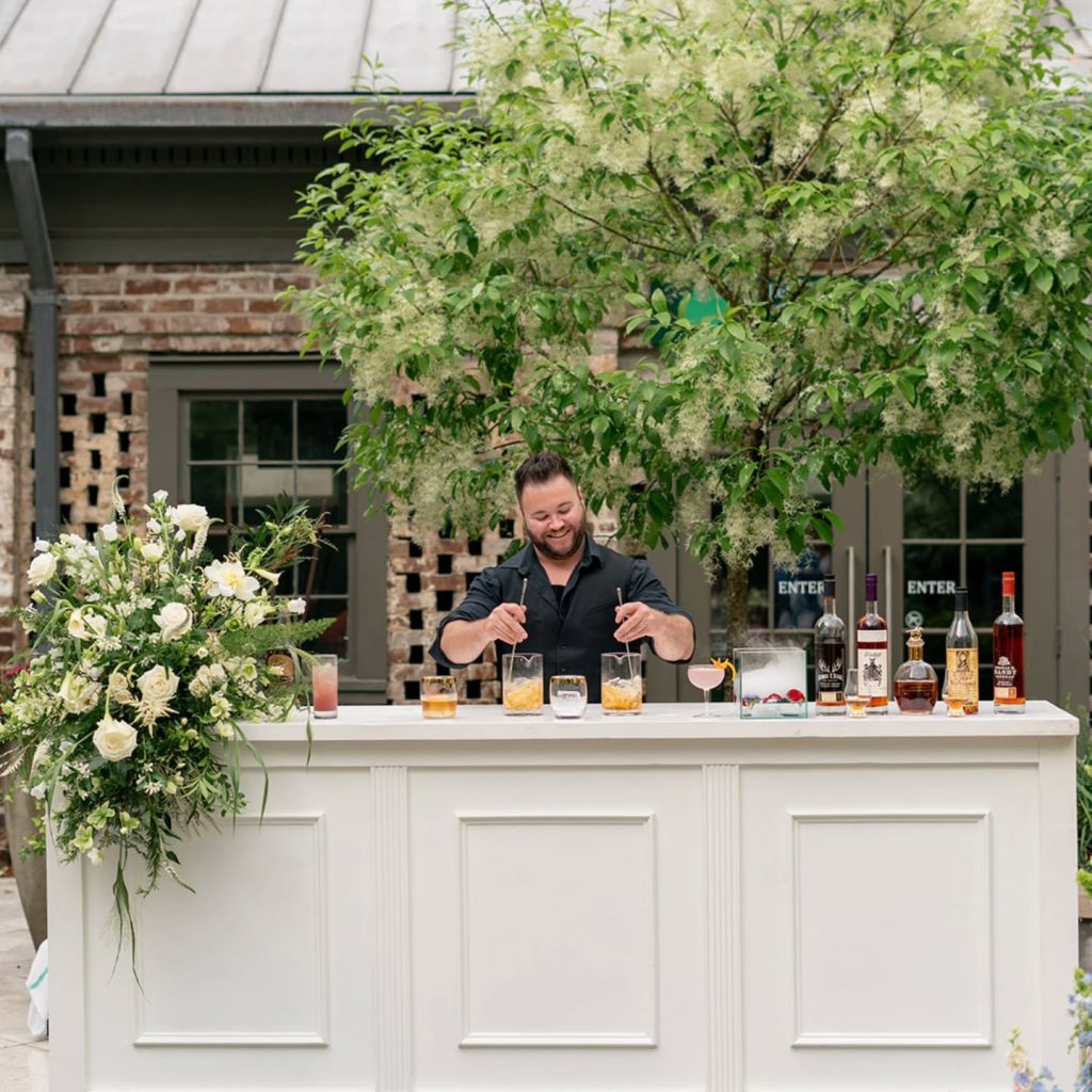 Bartender preparing cocktails at outdoor white bar decorated with white roses and greenery