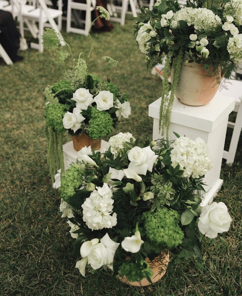 White and green floral arrangements in rustic urns displayed on tiered white pedestals at outdoor ceremony