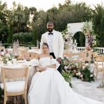 Bride in voluminous gown seated beside groom at outdoor reception with floral arch backdrop