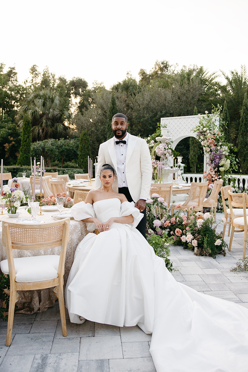 Bride in voluminous gown seated beside groom at outdoor reception with floral arch backdrop