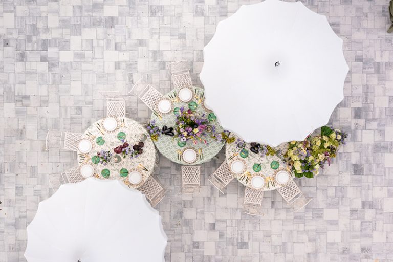 Overhead view of curved reception table arrangements with white umbrellas and floral centerpieces on gray stone patio