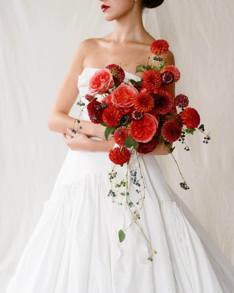 Bride in strapless white gown holding vibrant bouquet of coral and burgundy dahlias with cascading berries