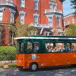 Old Town Trolley tour filled with passengers passing historic red brick Victorian mansion in Savannah