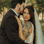 Bride with tattooed arms gazing at camera while groom kisses her forehead outdoors