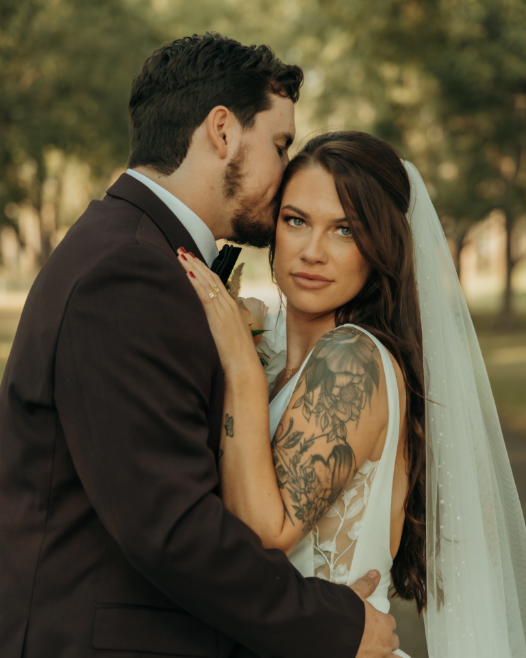 Bride with tattooed arms gazing at camera while groom kisses her forehead outdoors