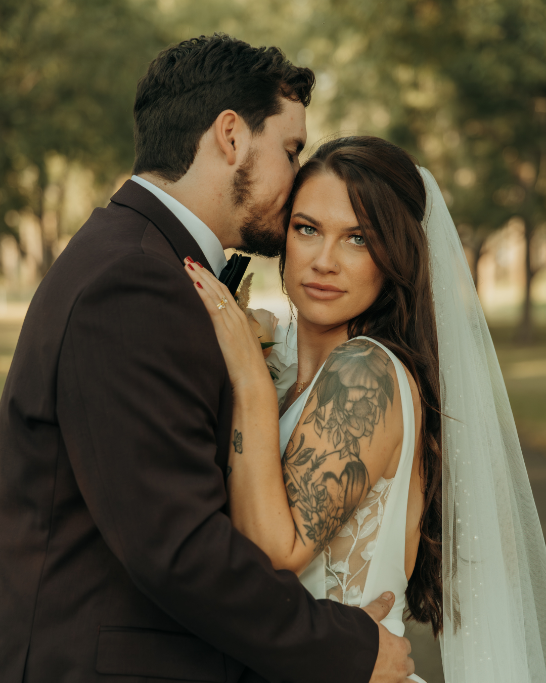 Bride with tattooed arms gazing at camera while groom kisses her forehead outdoors