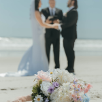 Pastel bridal bouquet on beach sand with couple and officiant during ceremony in background