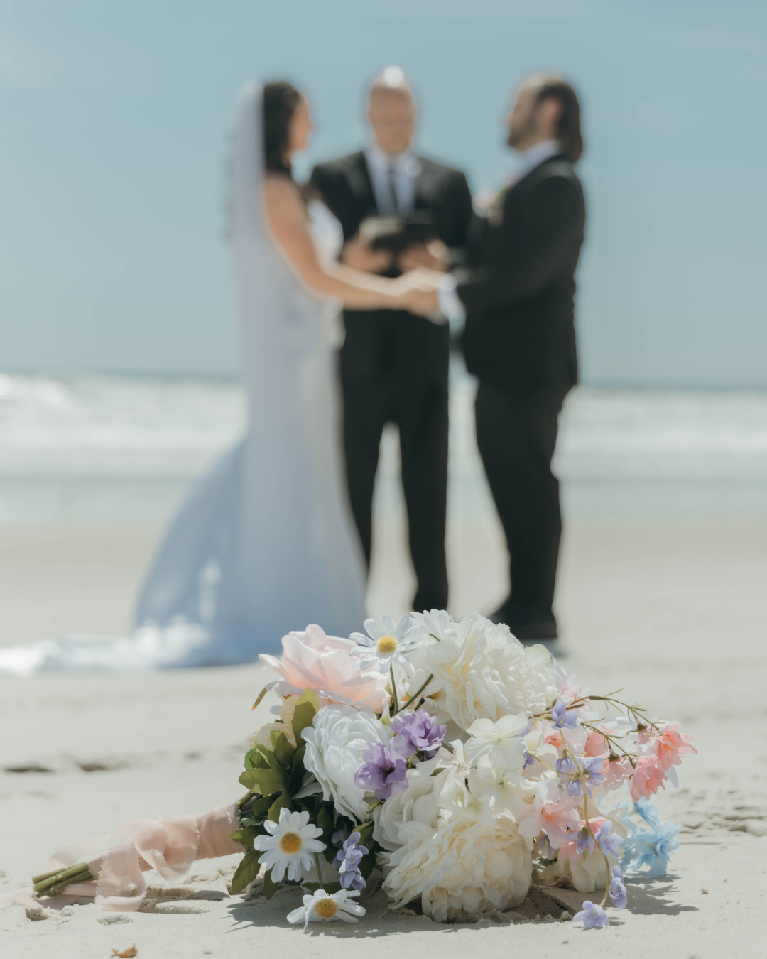 Pastel bridal bouquet on beach sand with couple and officiant during ceremony in background