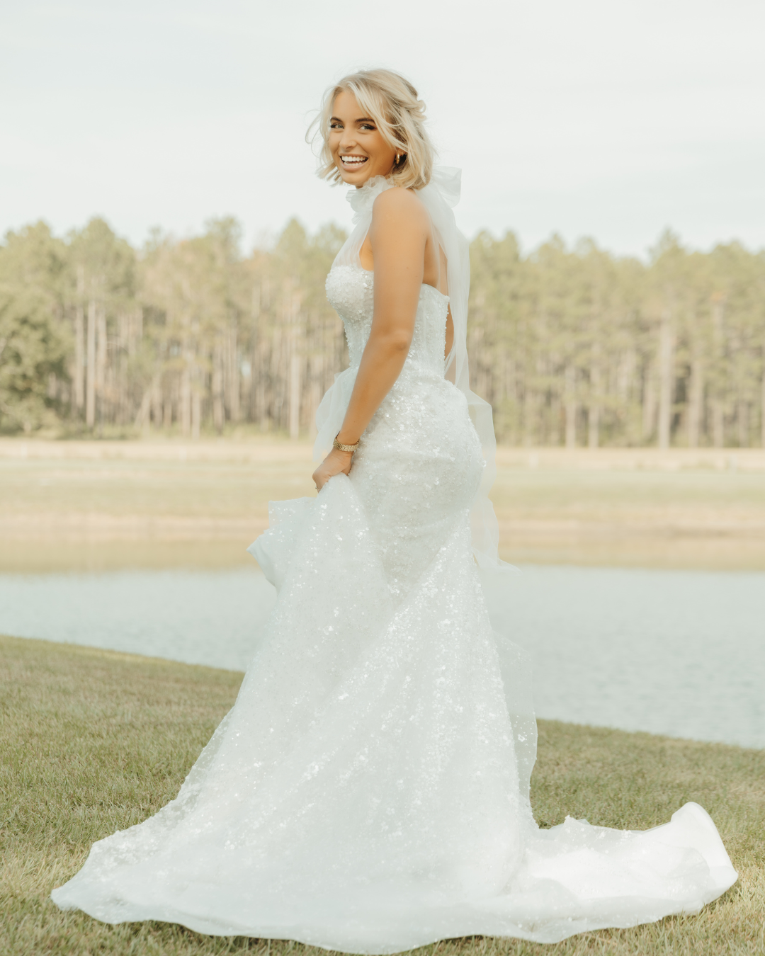 Bride in sparkly strapless gown with cathedral train twirling by lakeside with trees in background