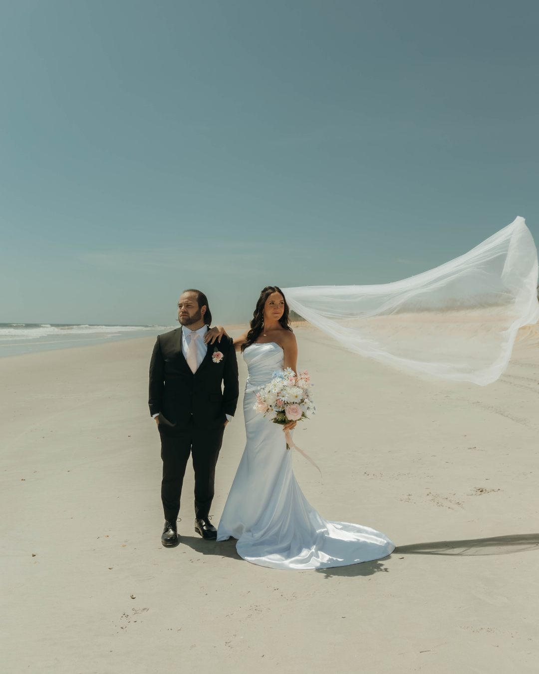 Newlyweds on coastal Georgia beach with bride's cathedral veil billowing in ocean breeze