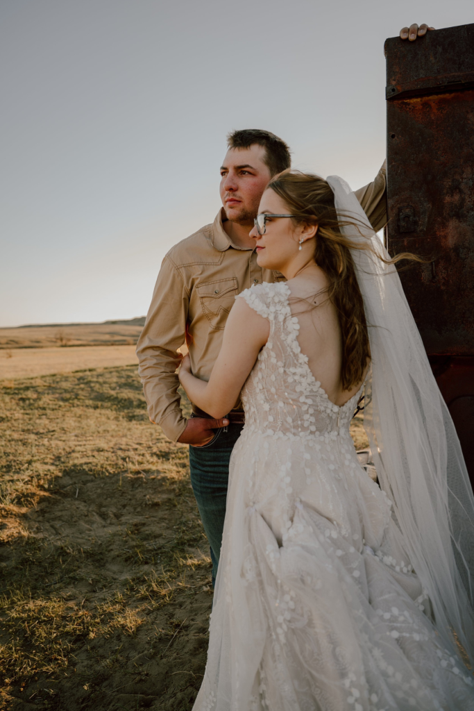 Bride and groom in casual western attire leaning against vintage farm equipment in open field at golden hour