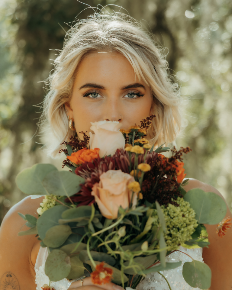 Bride with blonde updo holding autumn bouquet with burgundy mums, blush roses, and eucalyptus in outdoor setting