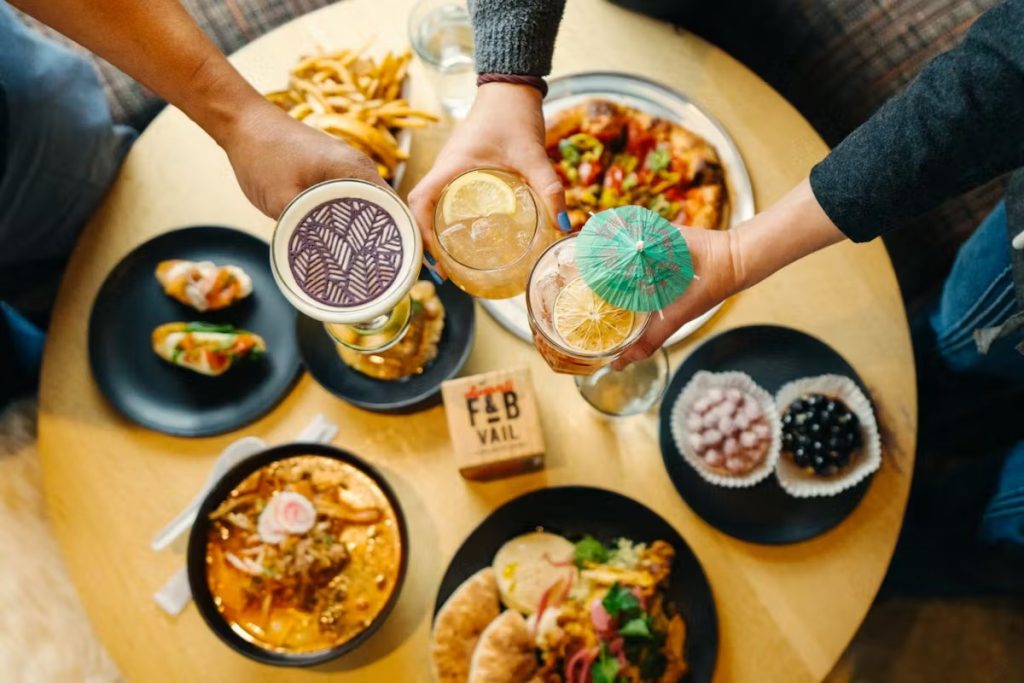 Overhead view of three hands toasting with craft cocktails over shared plates of diverse cuisine