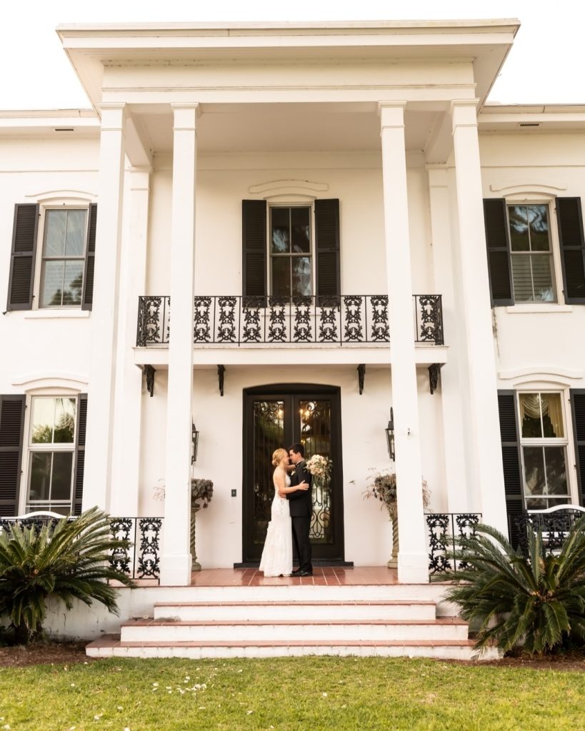 Bride and groom kissing in doorway of white antebellum mansion with black shutters and ornate ironwork balcony