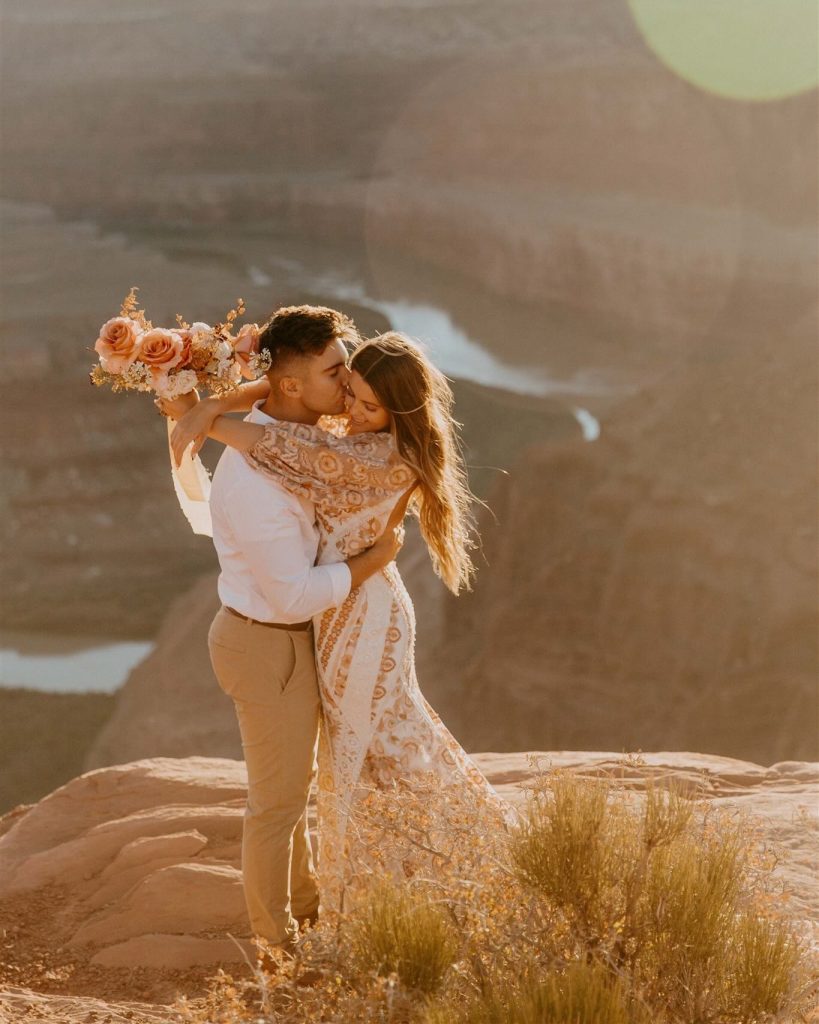 Couple embracing and kissing at golden hour on desert canyon overlook with bride holding peach and coral bouquet