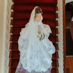 Bride in elegant white wedding dress and veil walks down stairs with red carpet runner