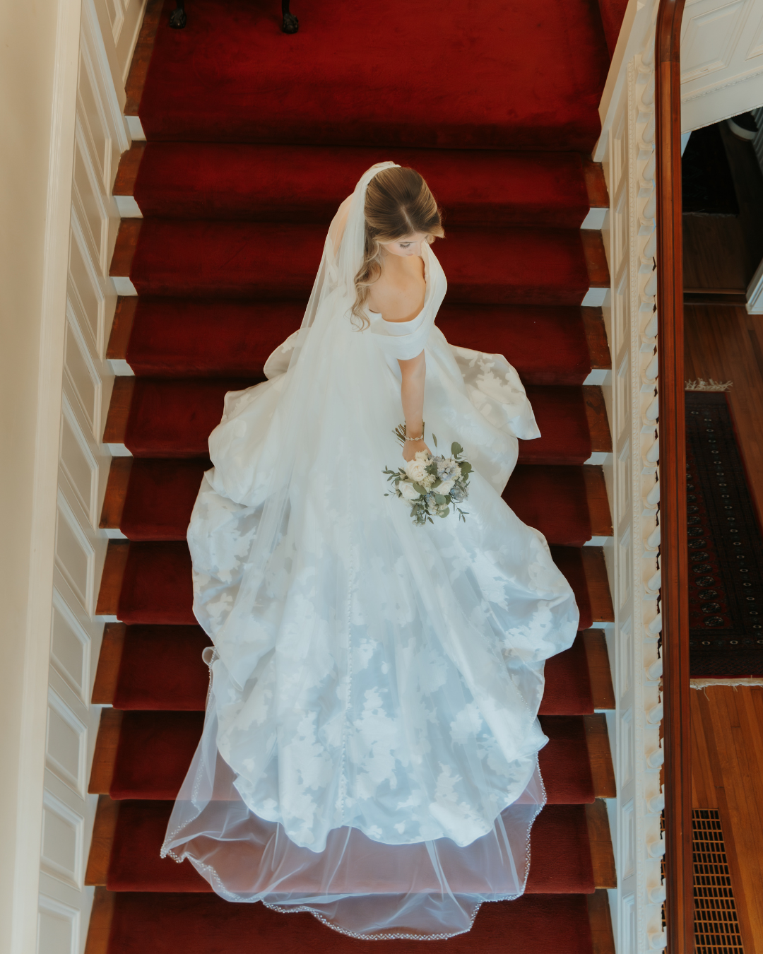 Bride in elegant white wedding dress and veil walks down stairs with red carpet runner