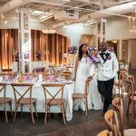Bride and groom walking through elegant wedding reception venue with purple floral arrangements and gold chandeliers