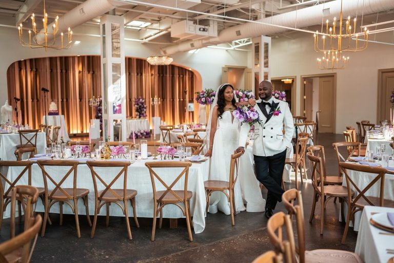 Bride and groom walking through elegant wedding reception venue with purple floral arrangements and gold chandeliers