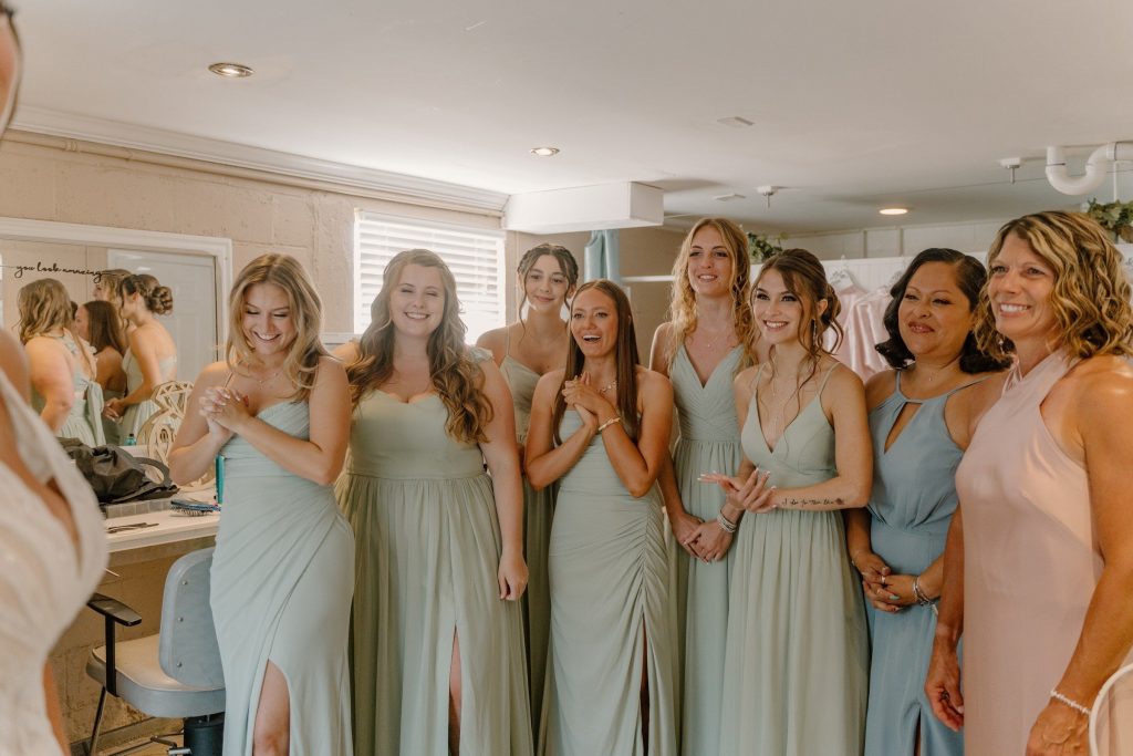Bridesmaids in sage green and blush gowns pose together during wedding preparation in elegant bridal suite