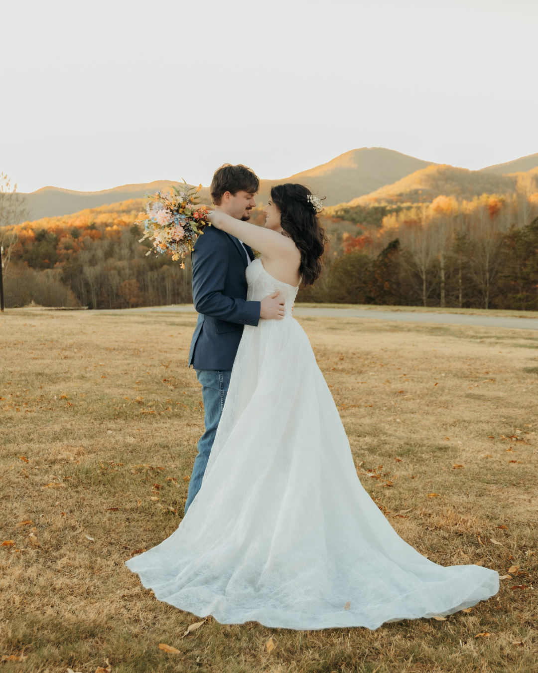 Newlyweds embracing in open field with autumn mountain foliage and golden hour lighting in background