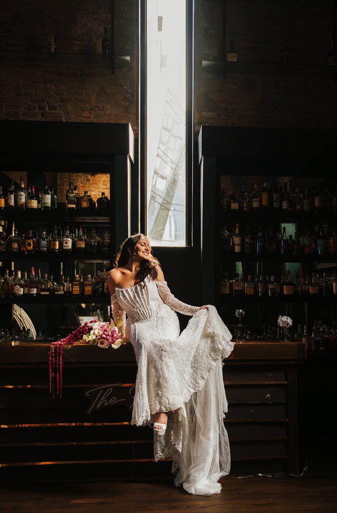 Bride in bohemian lace gown sits on bar counter in moody industrial venue
