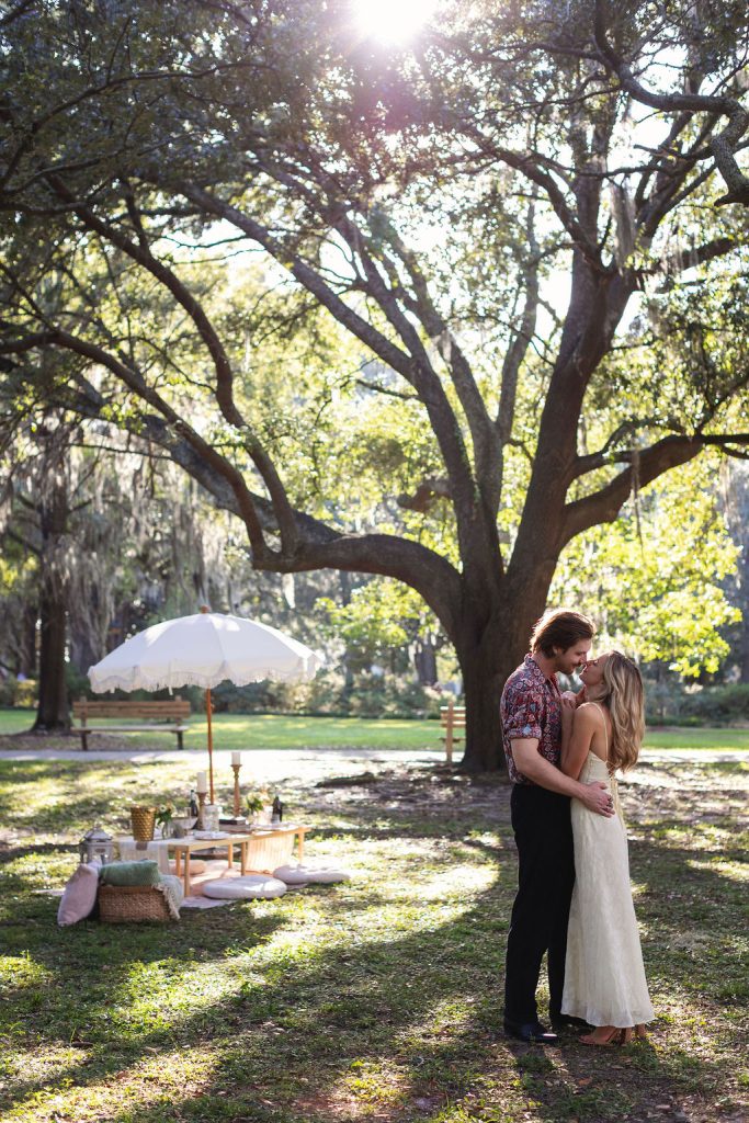 Couple embracing under a majestic oak tree with intimate picnic setup featuring white umbrella and cushions in Savannah