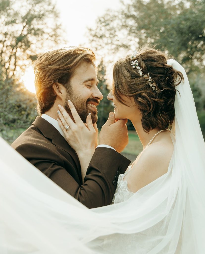 Bride touching groom's face as they smile at each other in golden hour outdoor portrait with flowing veil
