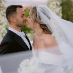 Newlyweds share intimate moment with flowing veil and white floral bouquet outdoors