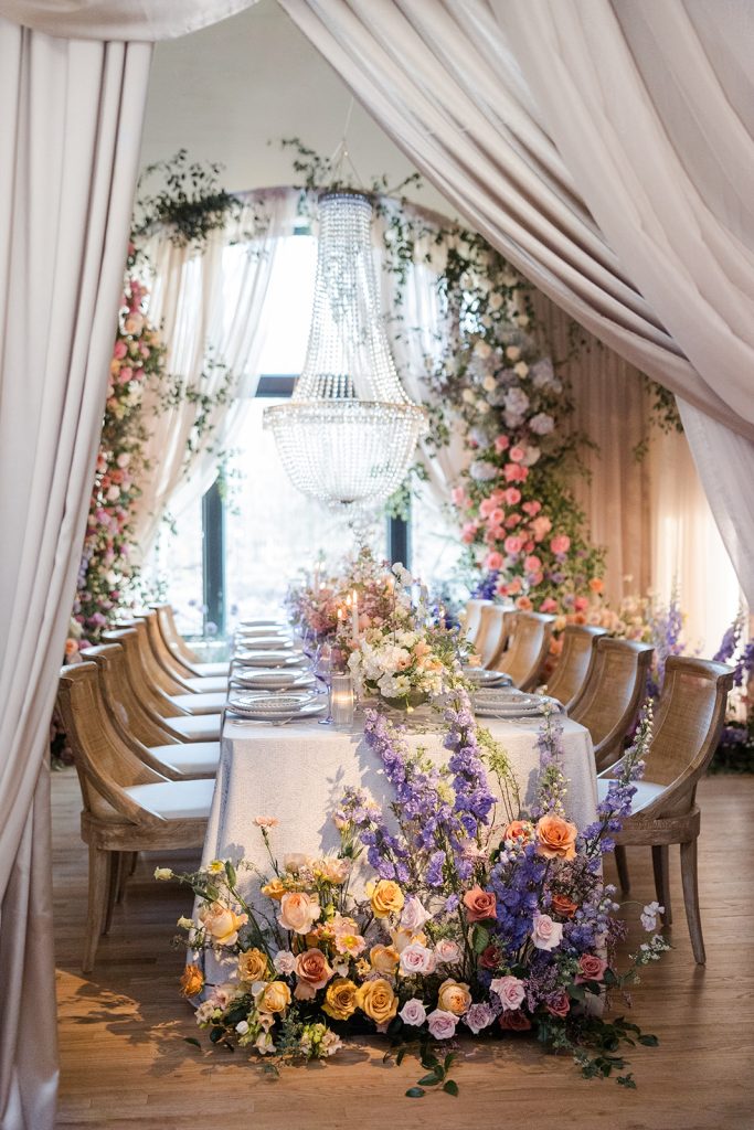 Wedding reception setup with draped fabric, chandelier, wooden chairs, and abundant floral arrangements