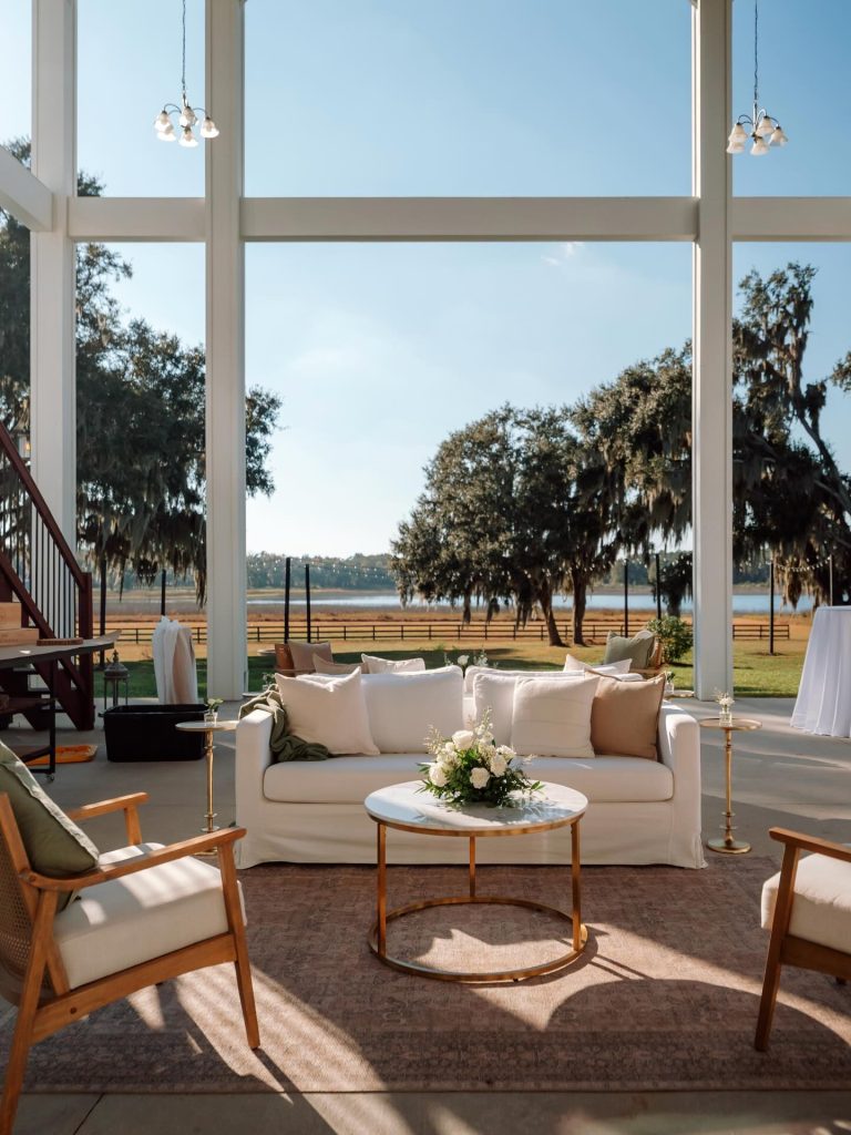 Elegant lounge area with white sofa and lakefront view framed by floor-to-ceiling windows and moss-draped oak trees