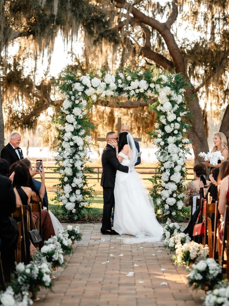 Outdoor wedding ceremony beneath Spanish moss-draped oak trees with white floral arch at The Barn at Covington Farm