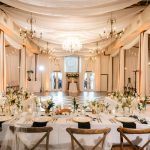 Elegant barn reception space with draped fabric ceiling, crystal chandeliers, string lights, and rustic wooden chairs