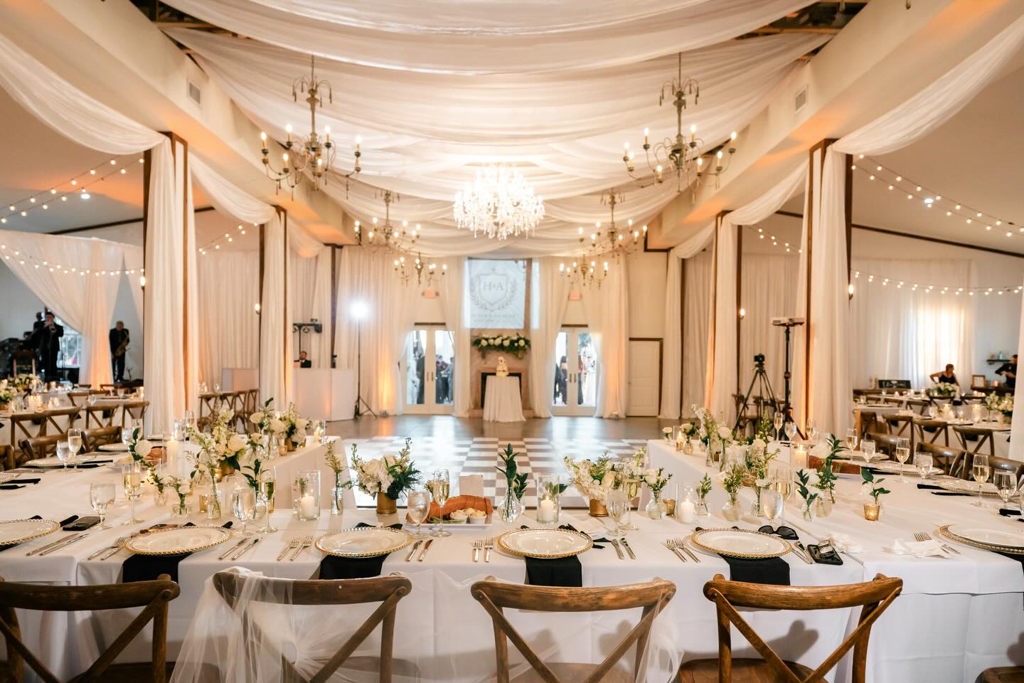 Elegant barn reception space with draped fabric ceiling, crystal chandeliers, string lights, and rustic wooden chairs