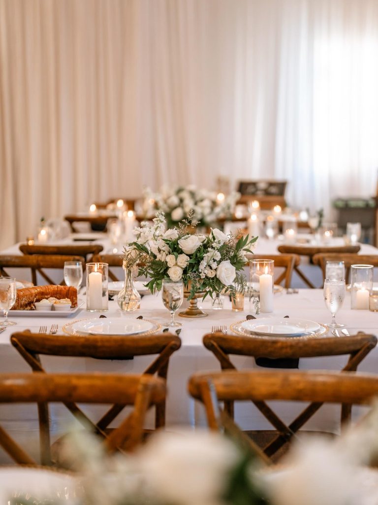 Elegant wedding reception table at The Barn at Covington Farm with white rose centerpiece, pillar candles, and rustic wood chairs against draped backdrop