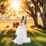 Bride in flowing white gown and veil posing under moss-draped oak trees at golden hour on farm property