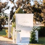 Elegant white welcome sign for Hector and Amanda's wedding at The Barn at Covington Farm, flanked by white floral arrangements and moss topiaries