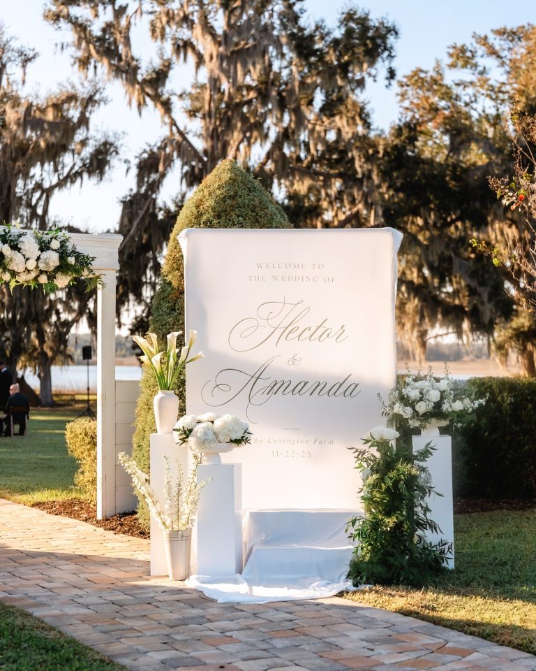 Elegant white welcome sign for Hector and Amanda's wedding at The Barn at Covington Farm, flanked by white floral arrangements and moss topiaries