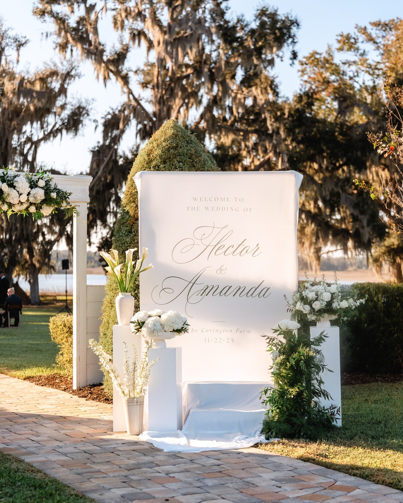 Elegant white welcome sign for Hector and Amanda's wedding at The Barn at Covington Farm, flanked by white floral arrangements and moss topiaries