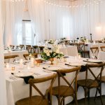 Elegant barn reception with white draping, string lights, wooden cross-back chairs, and gold-accented table settings