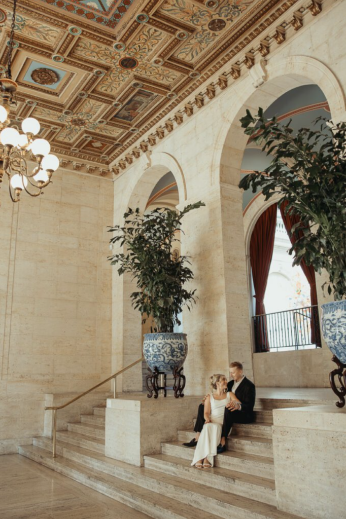 A couple sits together on grand marble steps inside the Columbus Museum of Art, surrounded by ornate painted ceilings, elegant archways, chandeliers, and large decorative planters.