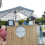 Bar Hop mobile bar with striped umbrella, pink balloon garland, and beverage dispensers at outdoor event