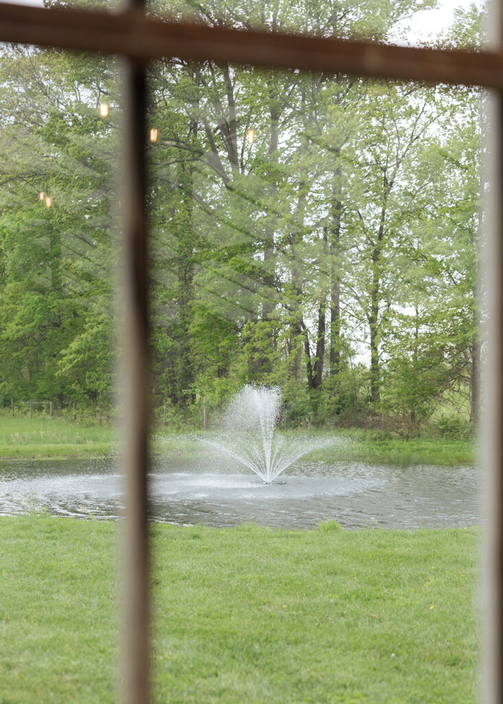 A peaceful pond with a spraying water fountain at Jorgensen Farms in Columbus, Ohio, surrounded by lush green grass and mature trees, viewed through a rustic window frame.