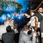 Bride and groom lifted on chairs holding pink cloth during hora dance at Savannah wedding reception with blue uplighting and greenery