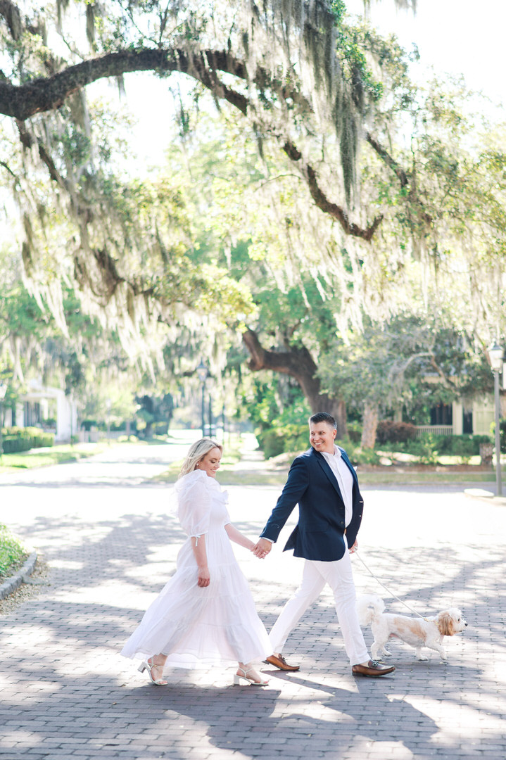 Couple walking hand-in-hand with small dog on cobblestone path beneath majestic live oak trees draped with Spanish moss