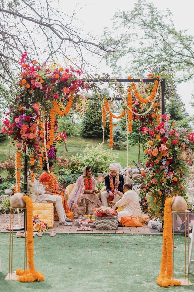 Colorful Indian wedding ceremony in Columbus with a floral mandap covered in vibrant pink, orange and red flowers, marigold garlands and lush greenery surrounding the couple during traditional wedding rituals.