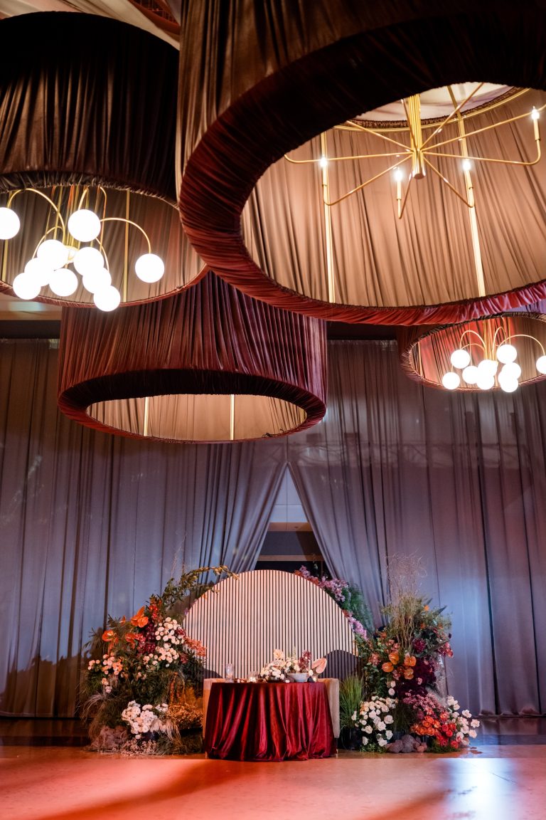 Wedding sweetheart table framed by pink and orange florals from Columbus florist Prema Designs, beneath oversized fabric chandeliers and dramatic draping.