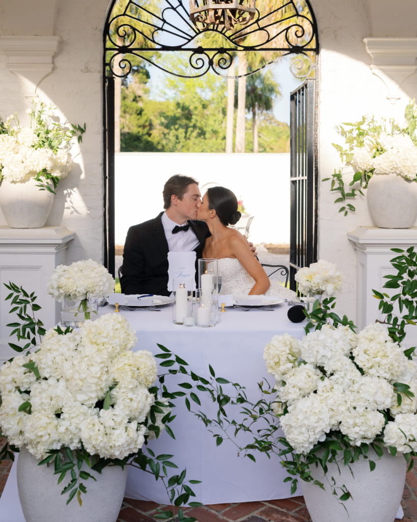 Bride and groom kissing at sweetheart table adorned with white hydrangeas and greenery at Jekyll Island wedding venue